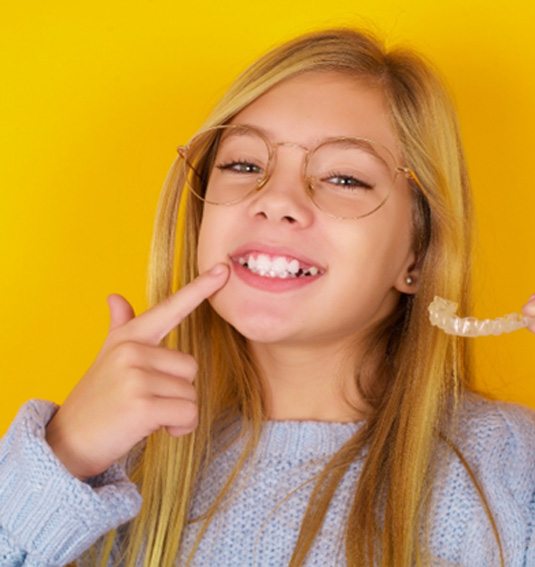 Young girl pointing at her smile and holding Invisalign aligners